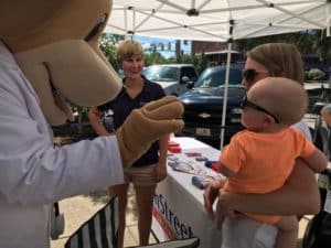 Mascot of MainStreet Family Urgent Care waves at a baby at the Eufaula Farmers Market