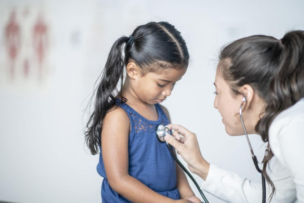 A young hispanic girl looks down at the stethoscope that her doctor is placing on her chest. Her doctor is a young caucasian woman and is wearing professional clothing.