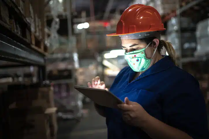Portrait of mid adult woman wearing face mask using digital tablet - working at warehouse / industry
