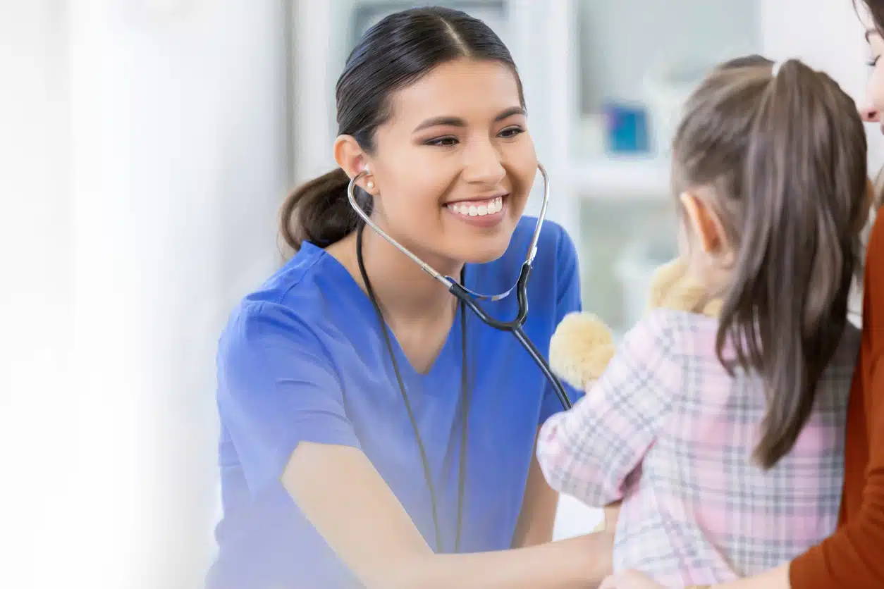 Confident female healthcare professional smiles as she uses a stethoscope during a young female patient's well check.